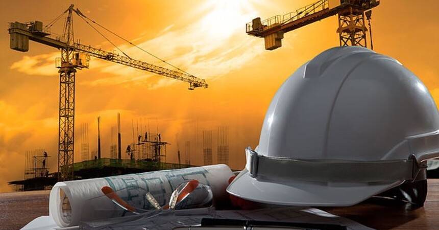 White safety helmet and rolled technical drawings on a construction site with tower cranes at sunset