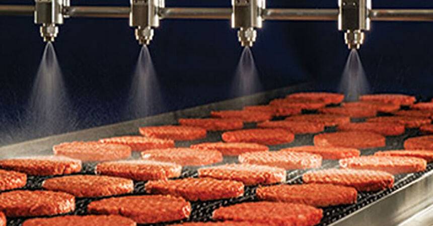 Burger patties moving along a stainless steel production line under spray nozzles