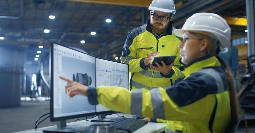 Two engineers in high-visibility jackets and hard hats reviewing a CAD model on screen in a factory